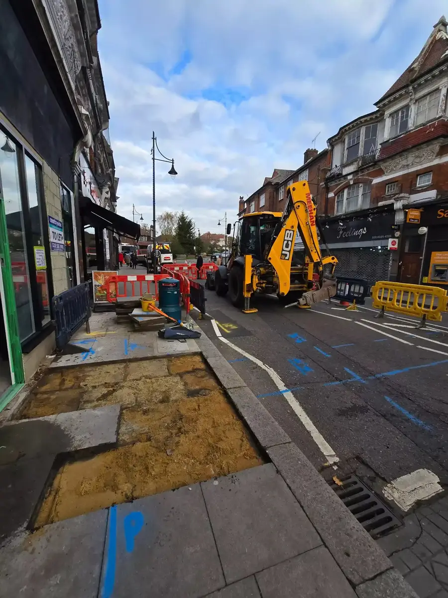 Excavator and safety barriers at street works on a high street, with a reinstated footpath trench in the foreground